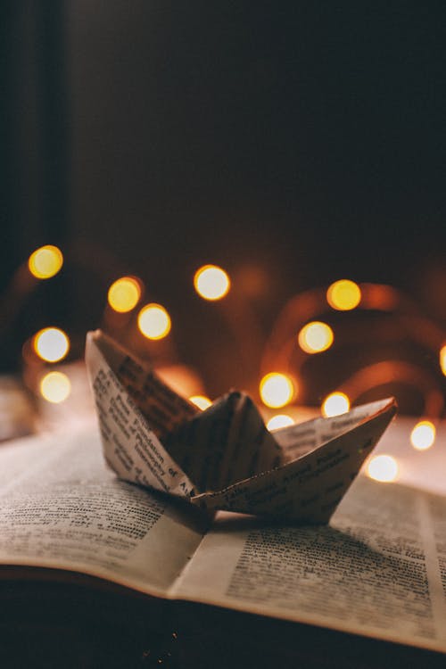 Paper ship made with book sheet placed on open book near glowing garland in bright room