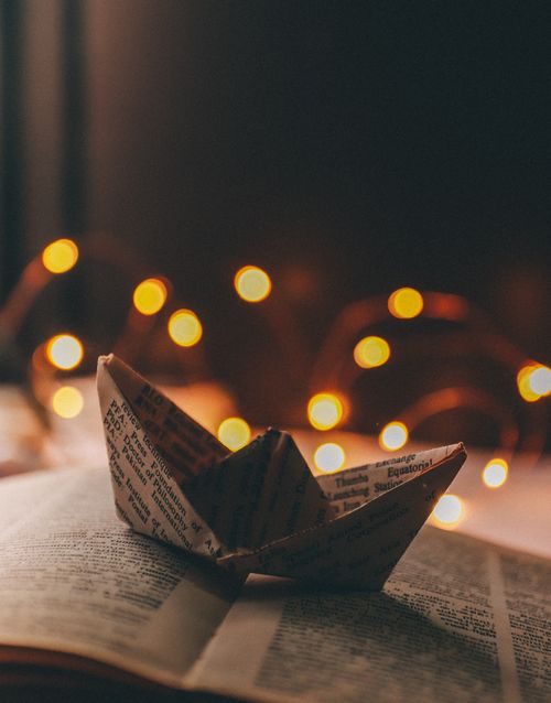 Free Origami ship made with book sheet placed on open book with illuminated garland on background in light room Stock Photo