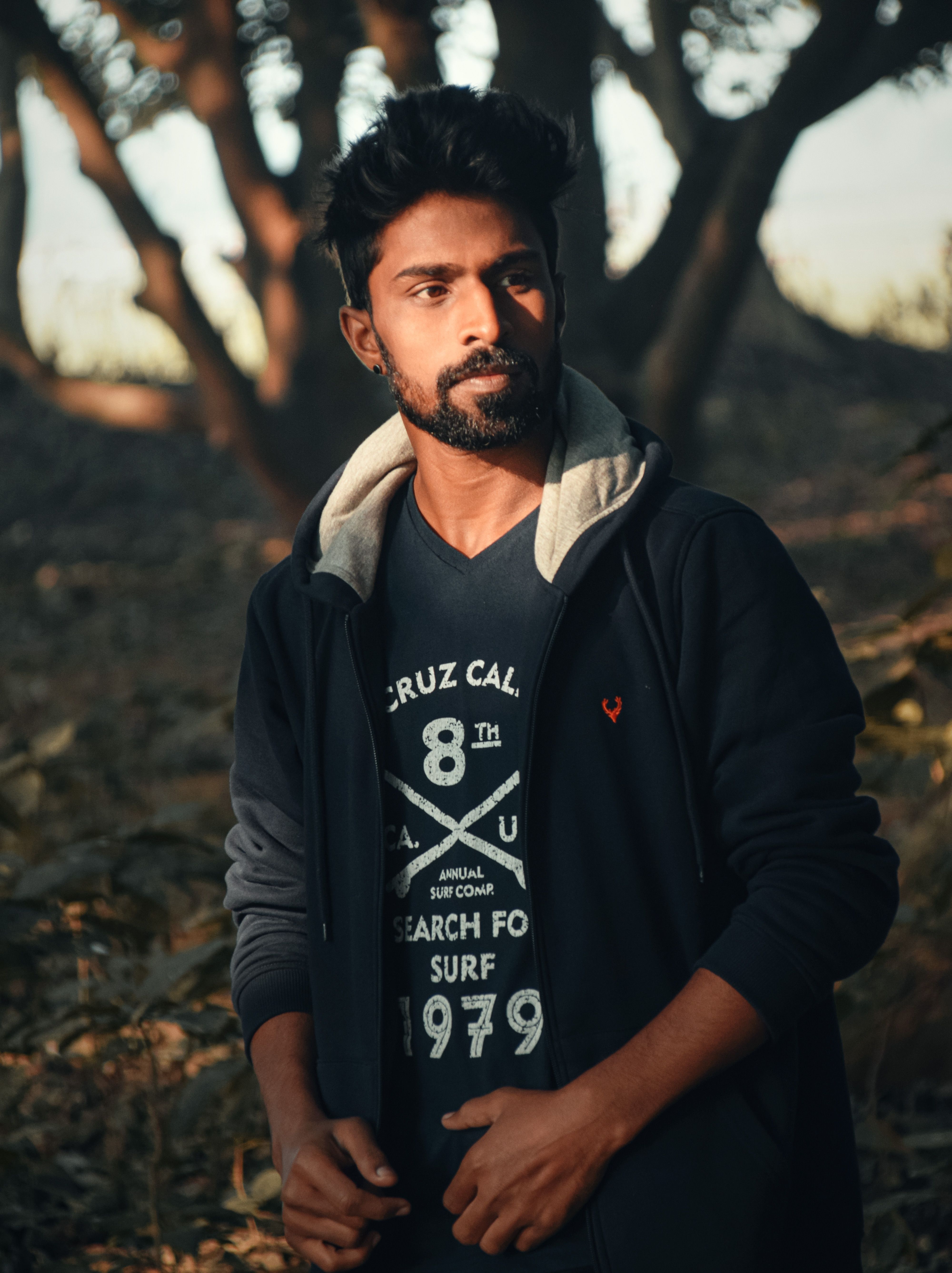 A contemplative man stands in a sunlit forest, offering a stylish portrait set in natural surroundings.