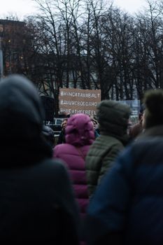 A group of people in winter clothing protests outdoors, holding a sign with hashtags.