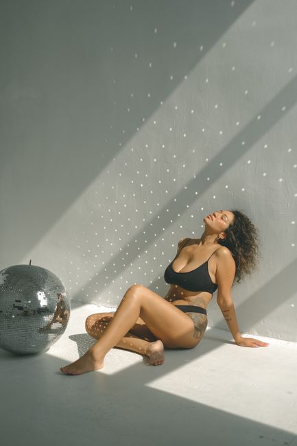 A woman in a bikini relaxes on the floor with a disco ball, basking in sunlight inside a minimalist setting.