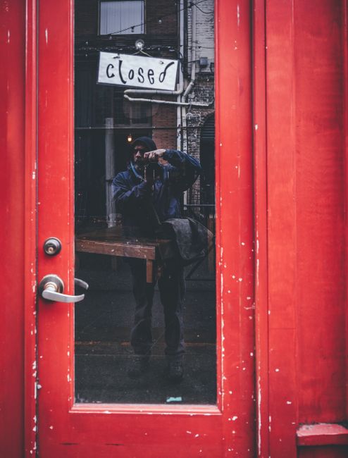 Full body of unrecognizable male traveler in warm casual clothes taking photo of aged red building with closed sign hanging on glass door