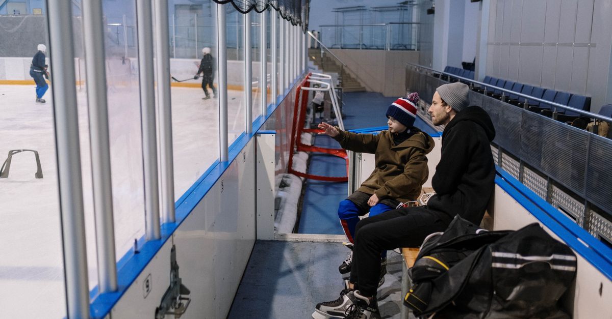 Father and son sitting on a bench at an ice skating rink, watching a game in progress.