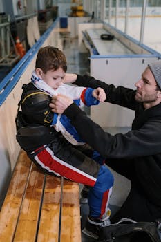 A father assists his young son with ice hockey gear in an indoor rink setting.