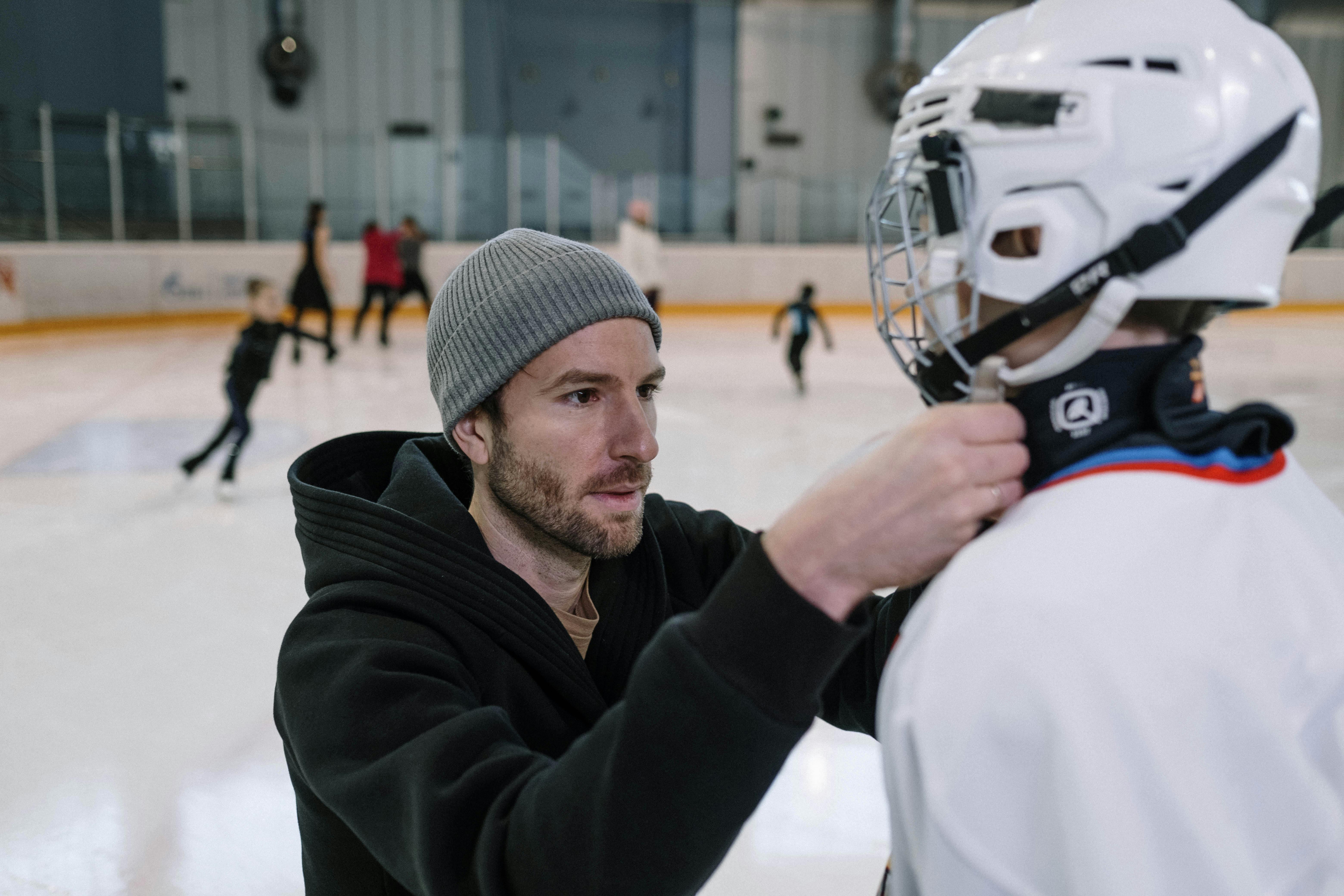 Father helps adjust son's hockey gear at an indoor ice rink, pre-game preparation.