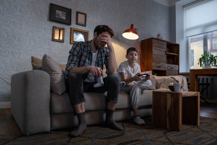 A Man And A Kid Playing Video Games While Sitting On The Sofa