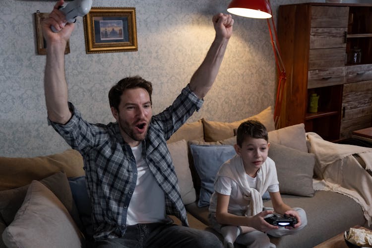 A Man And A Kid Playing Video Games While Sitting On The Sofa