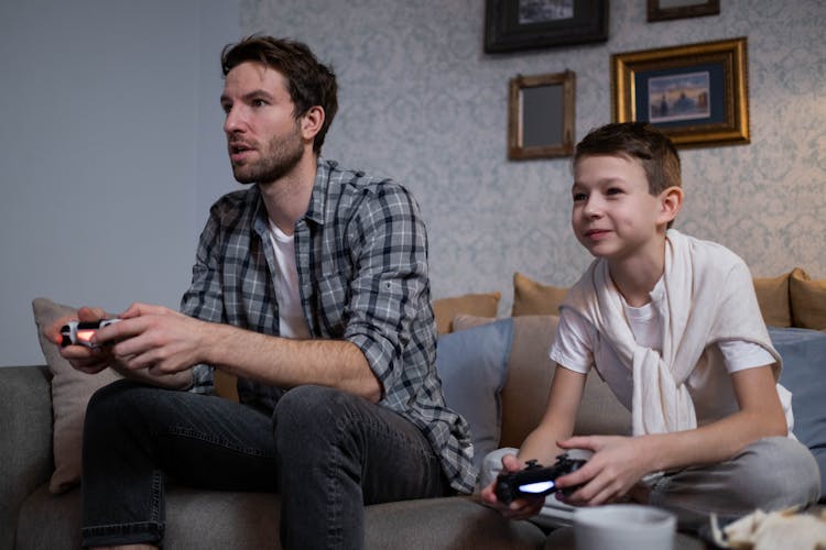 A Man And A Kid Playing Video Games While Sitting On The Sofa