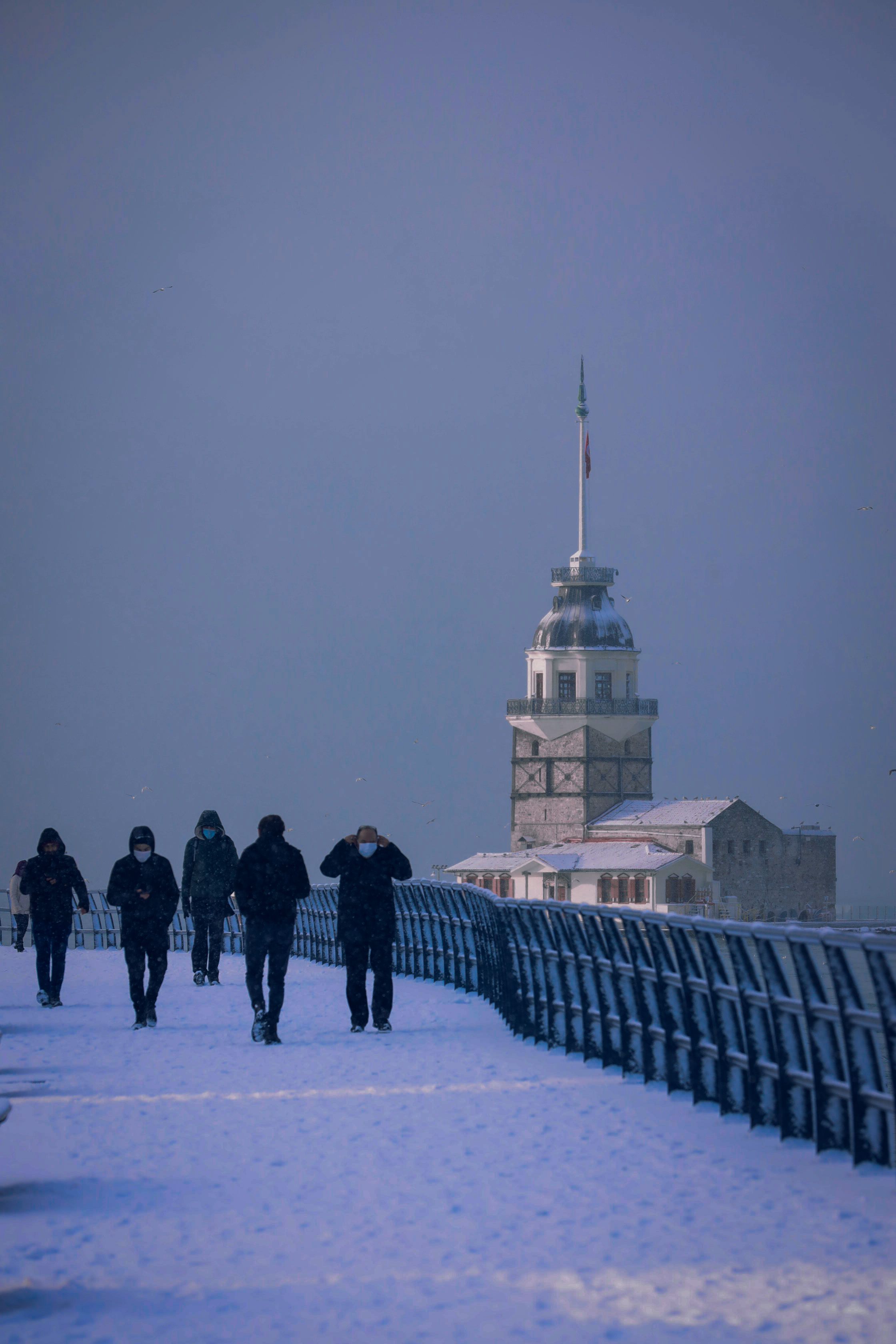 People walk on a snowy path with a historic lighthouse in the background, creating a serene winter scene.