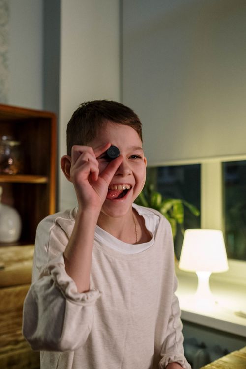 Free Joyful boy holding an object to his eye, smiling brightly in a warm, indoor environment. Stock Photo