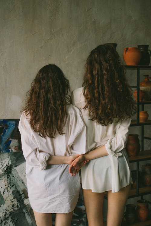 Back view of anonymous ladies wearing white shirts while standing in light studio near painting and shelves with pottery while holding hands