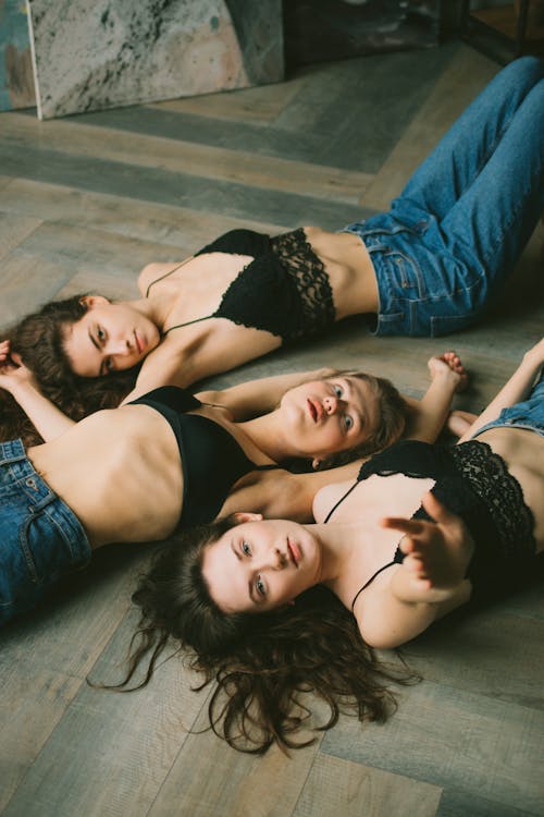 High angle of serious young female friends wearing black bras and jeans while lying on floor in bright studio near paintings and looking at camera
