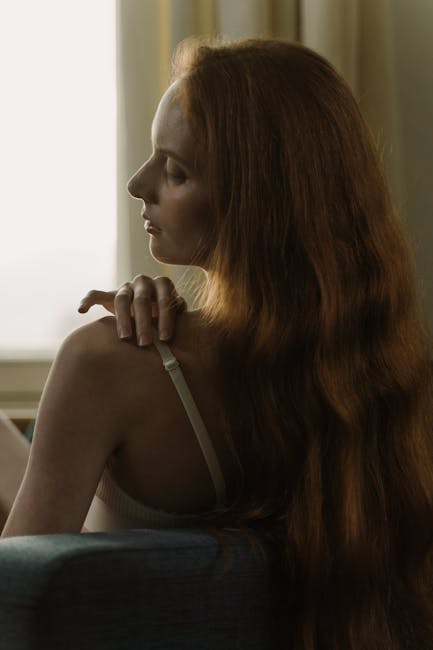 Sensual portrait of a woman with long red hair sitting by a soft-lit window.