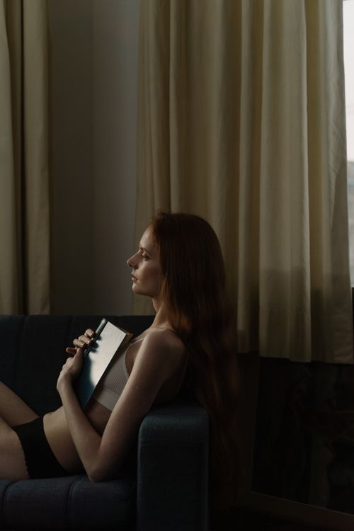 Free A woman in lingerie sits indoors, holding a book, relaxing on a couch. Stock Photo