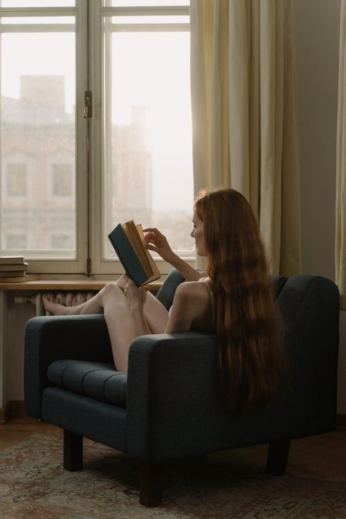 Free A woman reading a book in a cozy armchair by a window, enjoying a peaceful afternoon indoors. Stock Photo