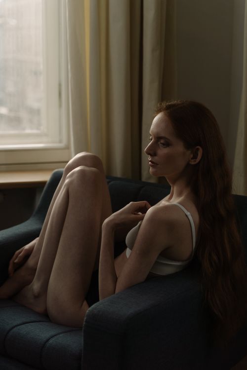 Free Redheaded woman in white top sitting on chair, lost in thought. Indoor portrait. Stock Photo