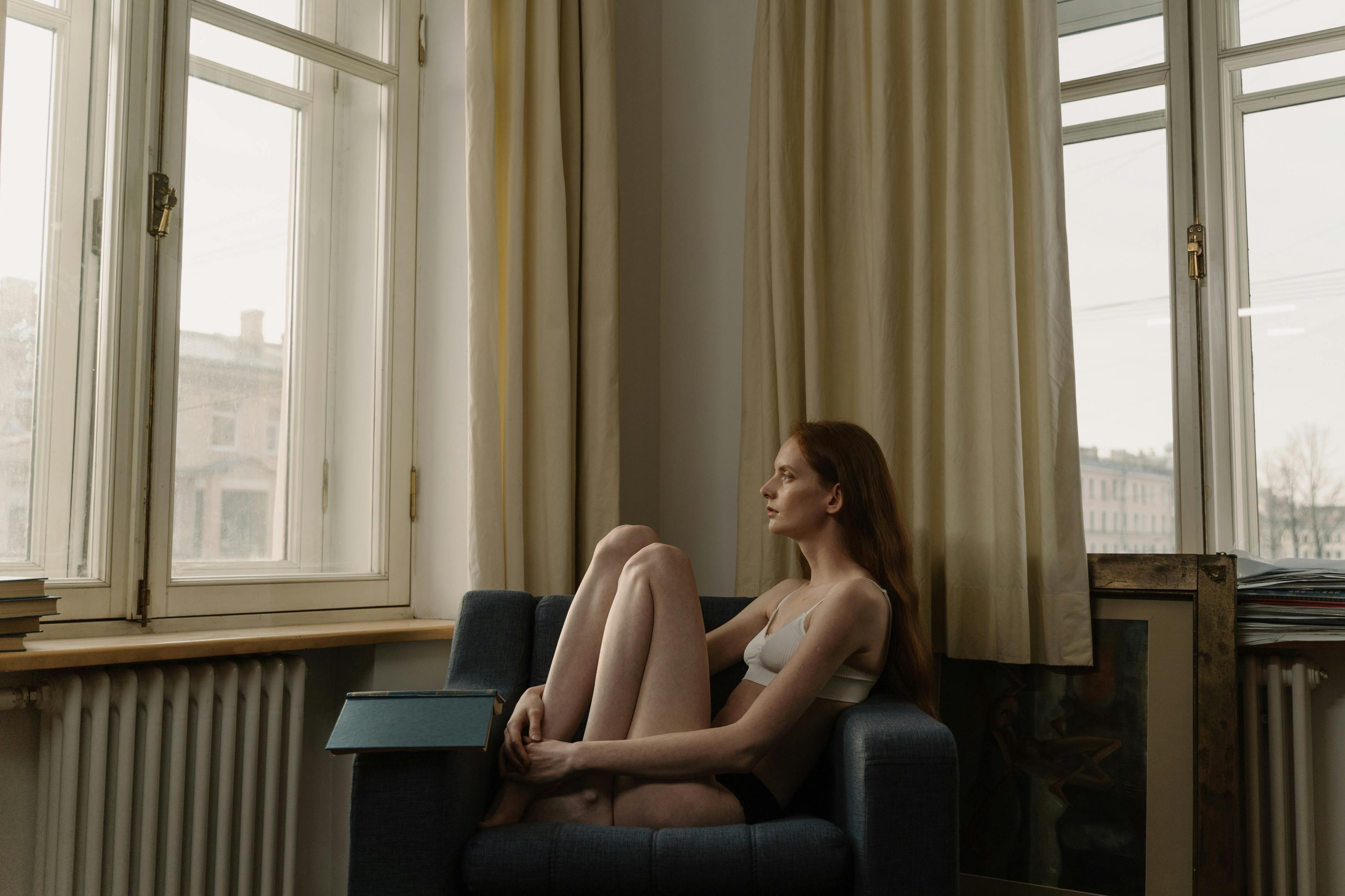 Free A woman in underwear sits in an armchair by large windows, embracing quiet introspection. Stock Photo