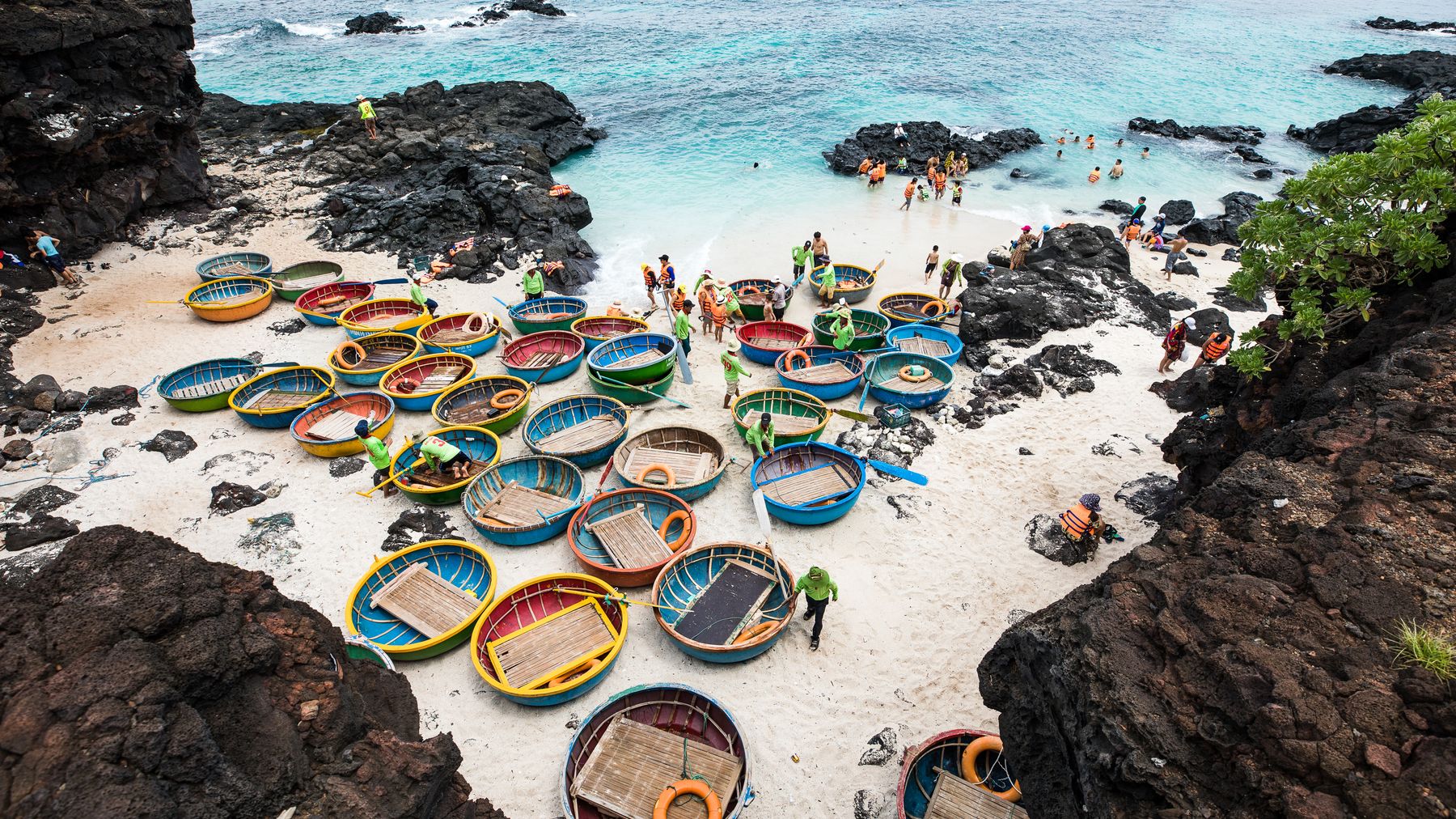 Aerial view of colorful basket boats by a rocky beach in Vietnam, with people enjoying the scene.