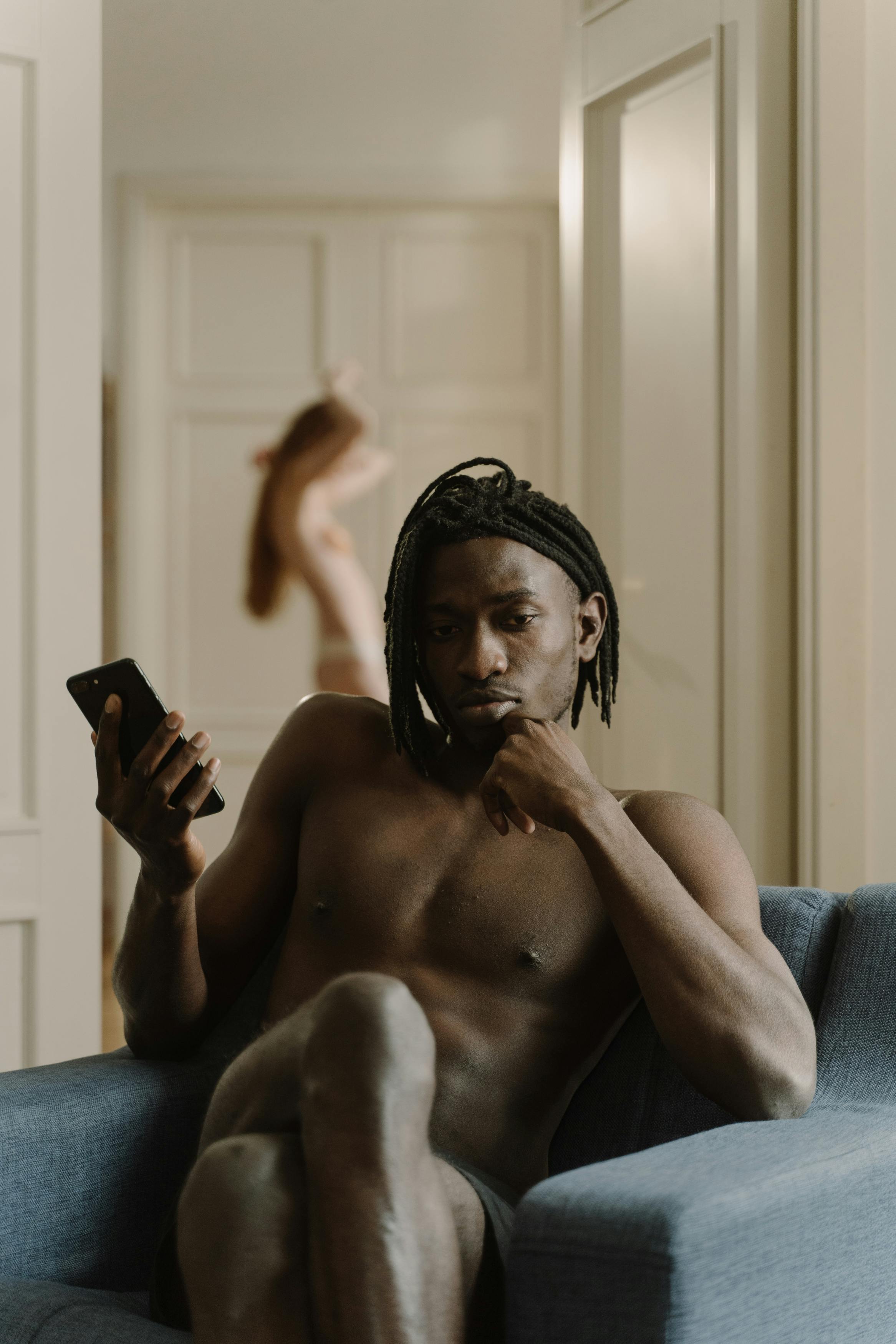 a shirtless man sitting on an armchair holding a smartphone