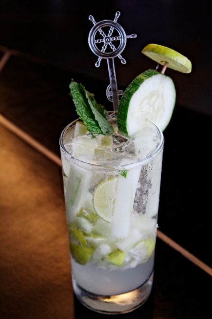 A tall glass of pale green cucumber lime juice garnished with a lime wheel and mint sprig, with whole cucumbers and limes on the counter beside it.