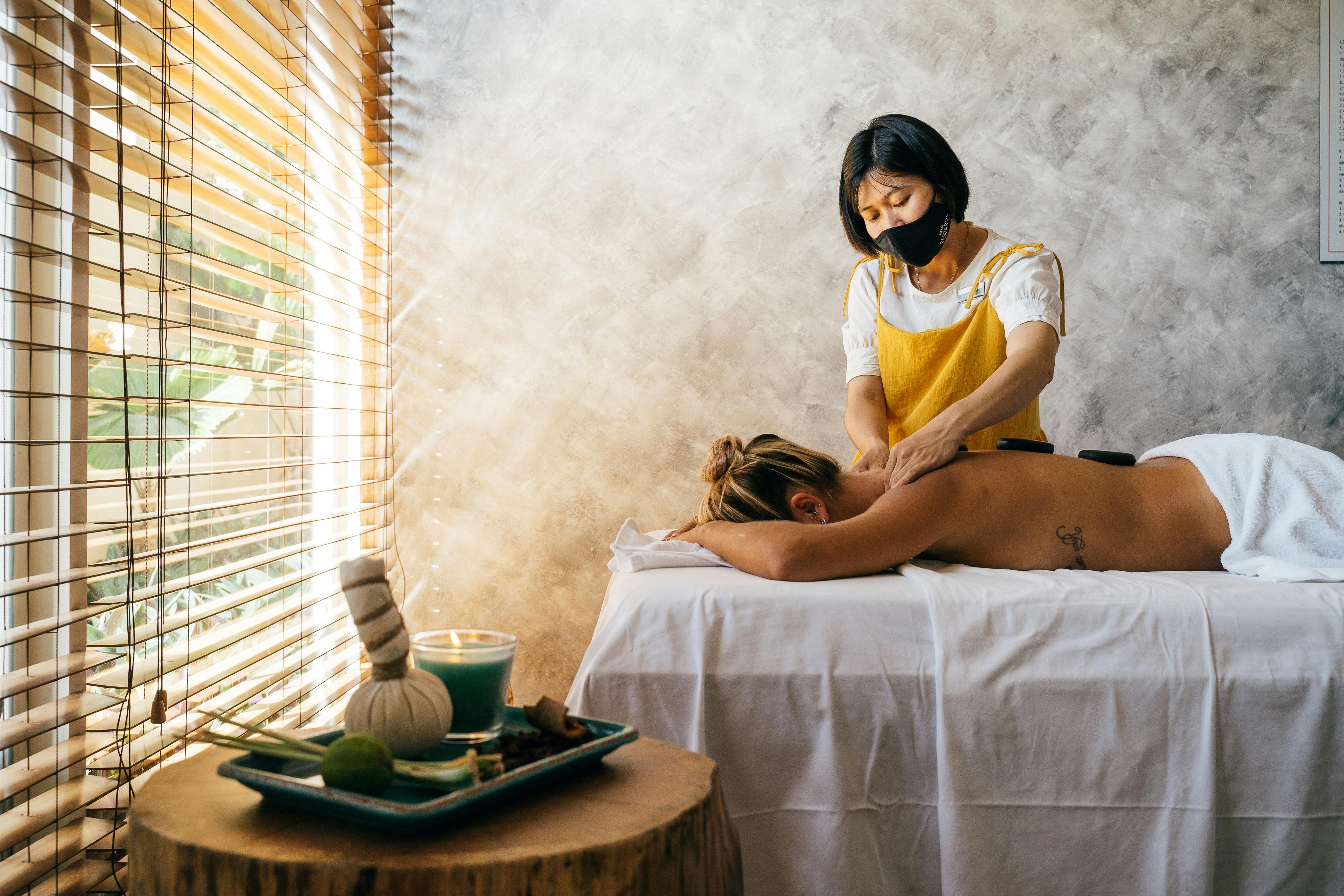 a woman wearing face mask massaging a topless woman lying down on a massage table