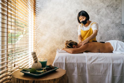 Free A Woman Wearing Face Mask Massaging a Topless Woman Lying Down on a Massage Table Stock Photo