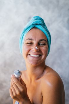 Adult woman smiling while applying skincare cream wearing a blue towel. Ideal for beauty and skincare themes.