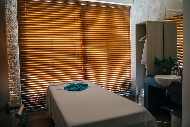 Treatment table with a folded teal towel in a room with wooden blinds and a corner basin