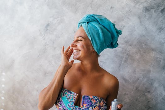 Happy woman with towel on head applying facial cream, embracing skincare routine indoors.