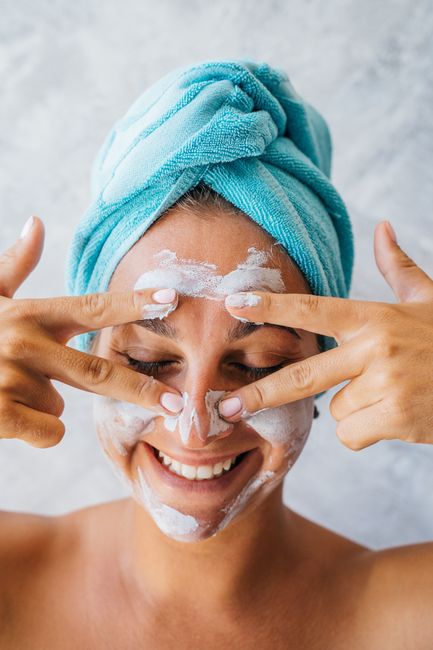Portrait of a smiling woman applying facial cream with towel wrap, showcasing a happy skincare routine.