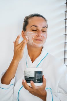 Woman in bathrobe applying moisturizer with eyes closed in a relaxing skincare routine.