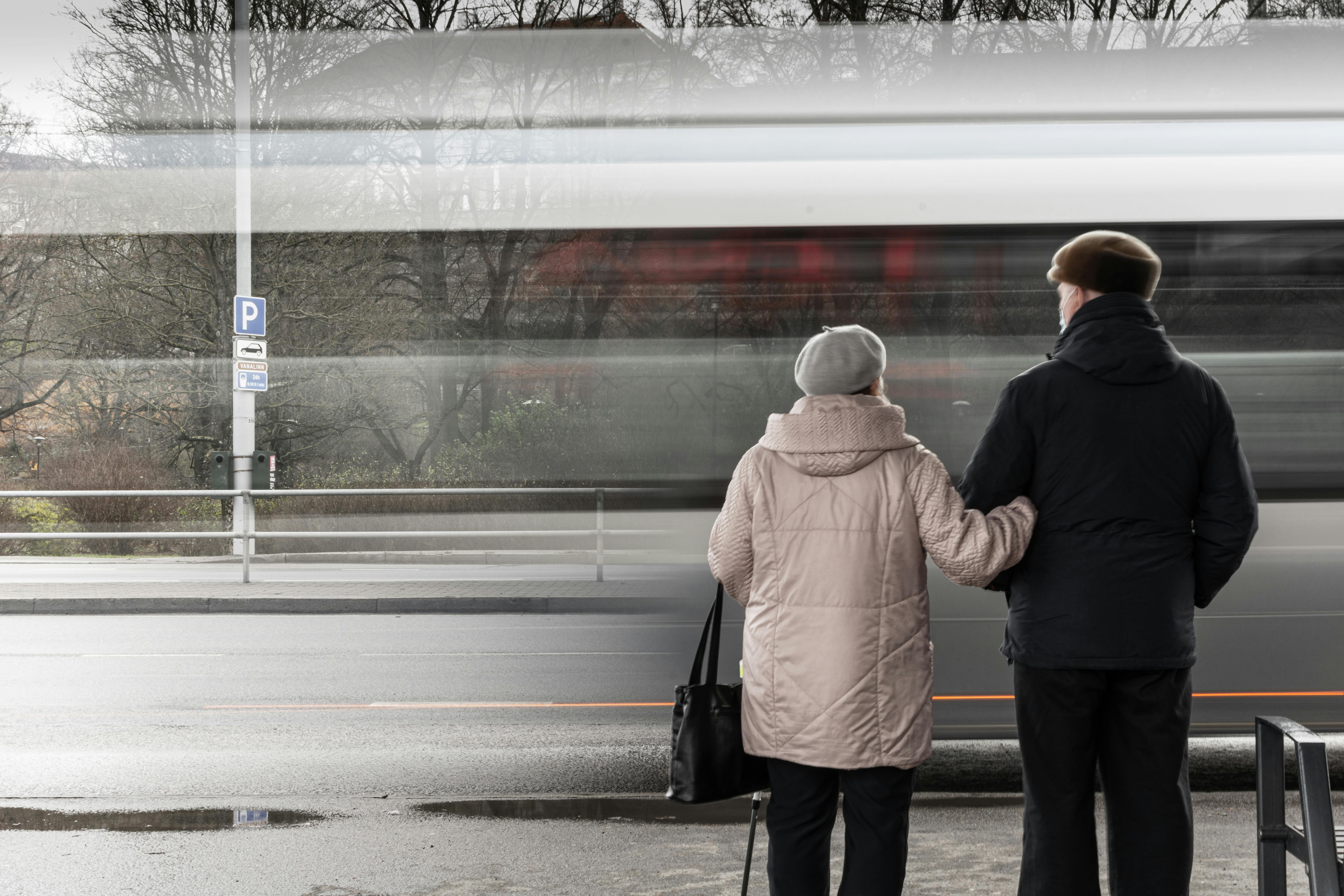 An elderly couple in jackets and hats hold hands, watching passing traffic at a bus stop.