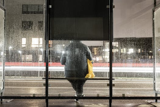 Person waiting at a rainy bus stop at night with light trails in the background.