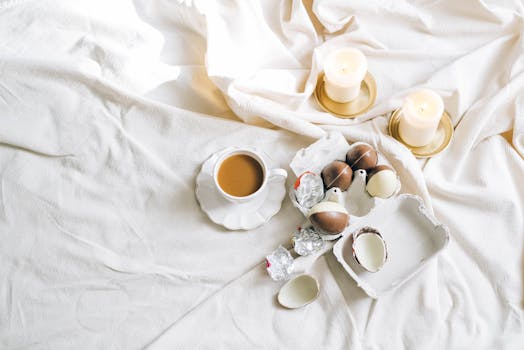 A cozy setting with a cup of coffee, chocolate eggs, and lit candles on a soft white fabric.