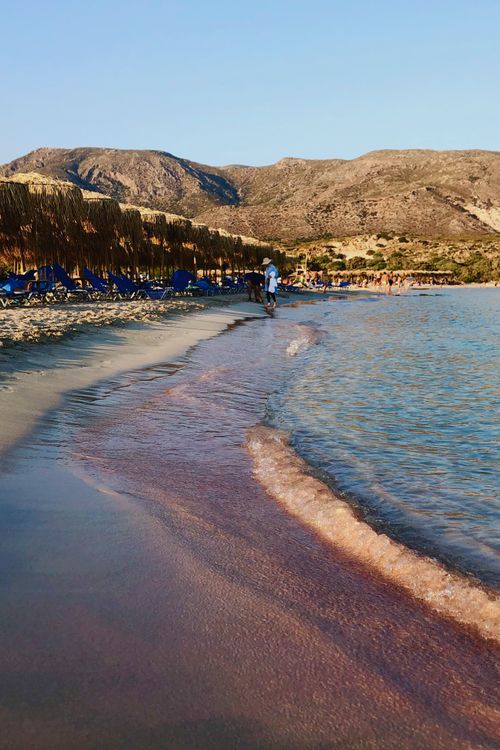 Free Serene beach scene featuring pink sand, clear blue waters, and distant mountains under a clear sky. Stock Photo