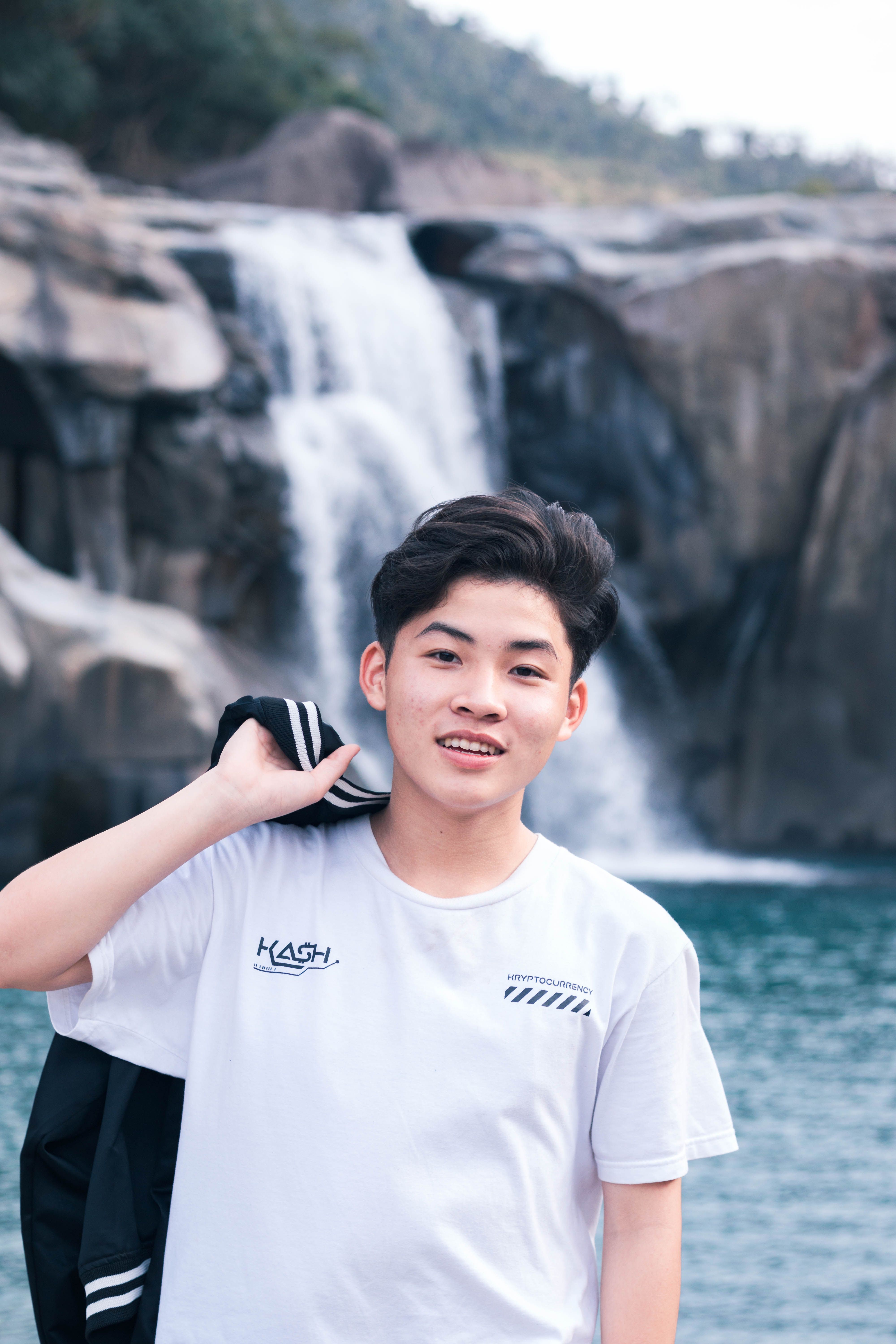 A young man in a white t-shirt smiles cheerfully in front of a waterfall.