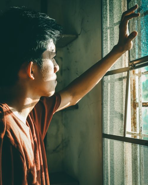 Free Young man stands thoughtfully by the window with sunlight and shadows creating a dramatic effect. Stock Photo
