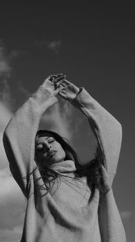 Elegant black and white portrait of a woman with eyes closed and arms raised against the sky.