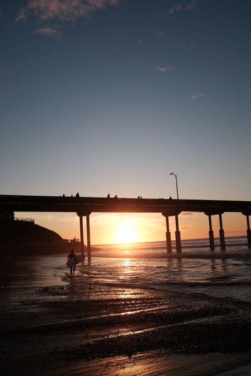 Free A lone surfer walks along the beach during a stunning sunset at Ocean Beach Pier in San Diego, California. Stock Photo
