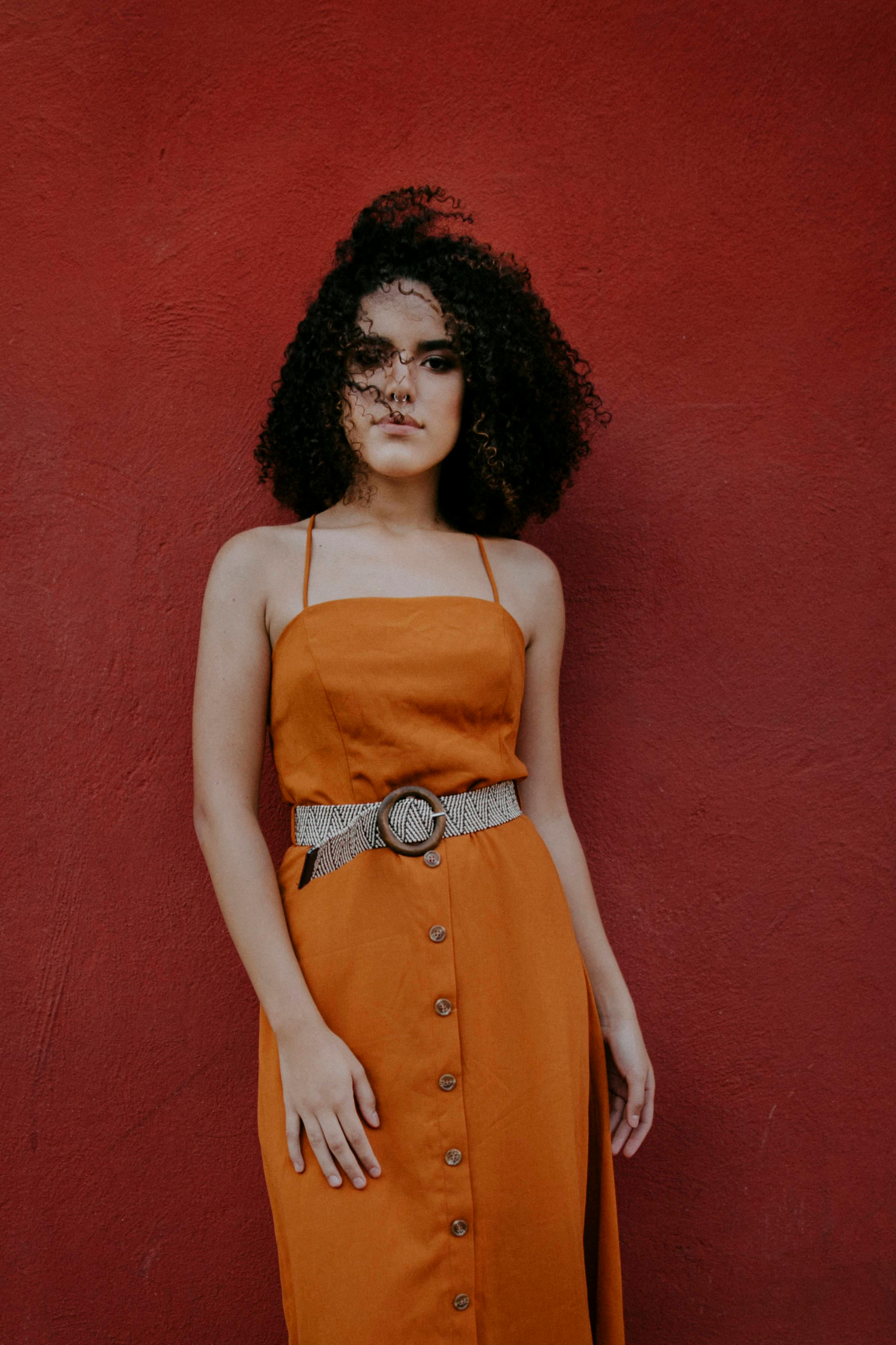 Woman with curly hair posing in an orange dress against a bold red background.
