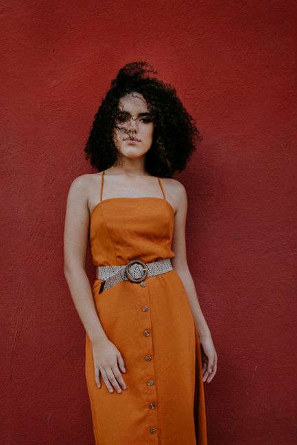 Woman with curly hair posing in an orange dress against a bold red background.