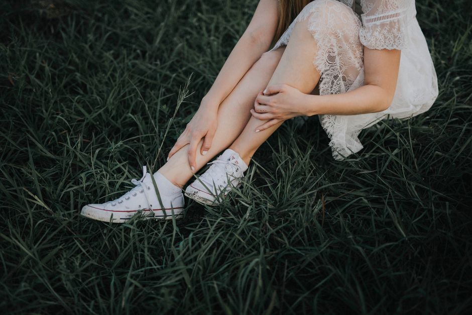 A woman in a white lace dress sitting on green grass wearing white sneakers.