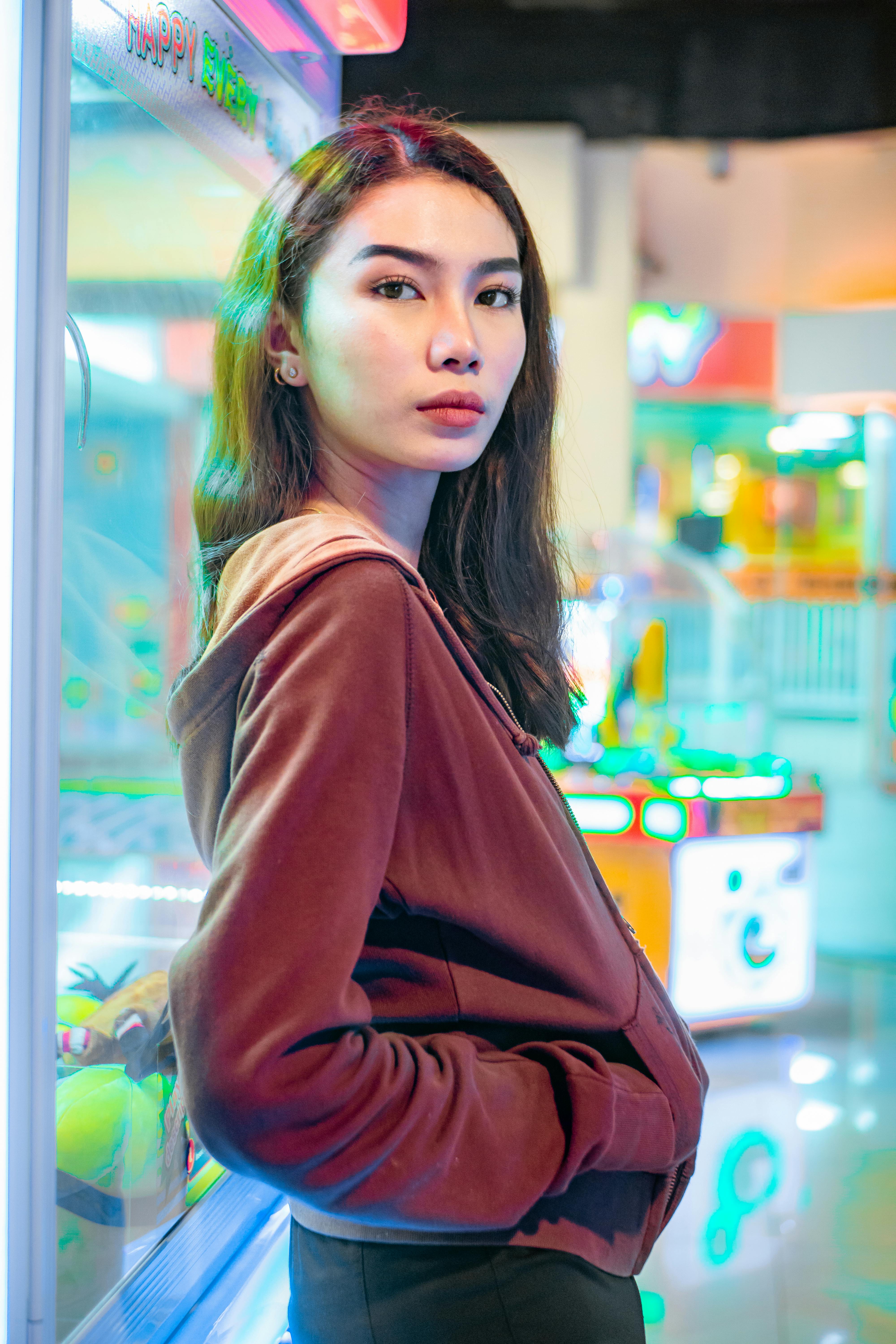 Serious woman wearing brown hoodie in a vibrant arcade setting, reflecting neon lights.