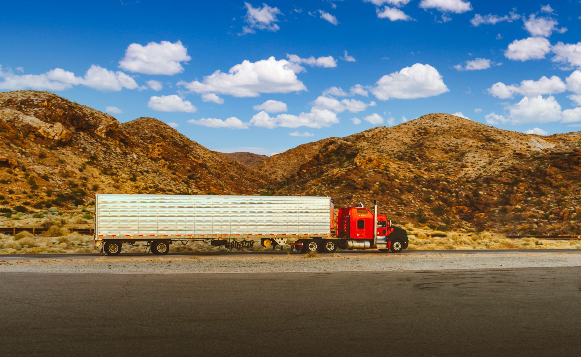 Red semi-truck with a white trailer on a desert highway below rocky hills