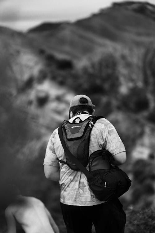 Black and white back view of anonymous male hiker with backpack wearing cap standing against rocky cliff in nature during trekking on blurred background