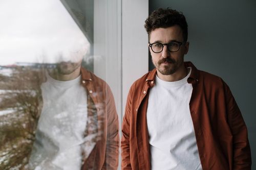 Free Portrait of a man with glasses standing by a window, looking thoughtful. Stock Photo