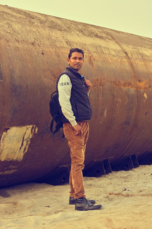 Man Standing Next to a Big Rusty Tube on a Beach 