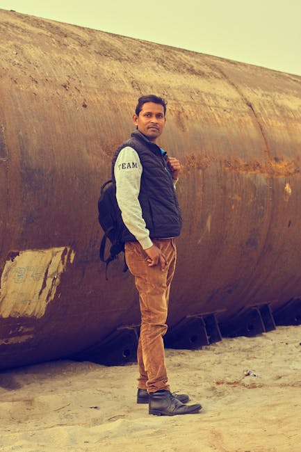 A man with a backpack stands beside a rusty pipe on a sandy beach. Adventure awaits.