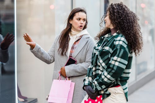 Multiethnic female friends with shopping bags looking at each other and communicating while standing on sidewalk near shop window on street