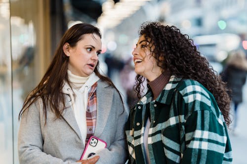 Optimistic multiracial female friends in outerwear chatting while standing near showcase of store on street in city on blurred background